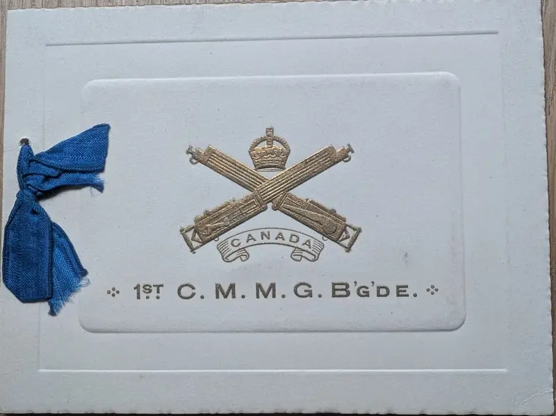 Embossed gilt card, crossed Vickers machine guns under a crown, '1st C.M.M.G. B'g'de', blue silk ribbon